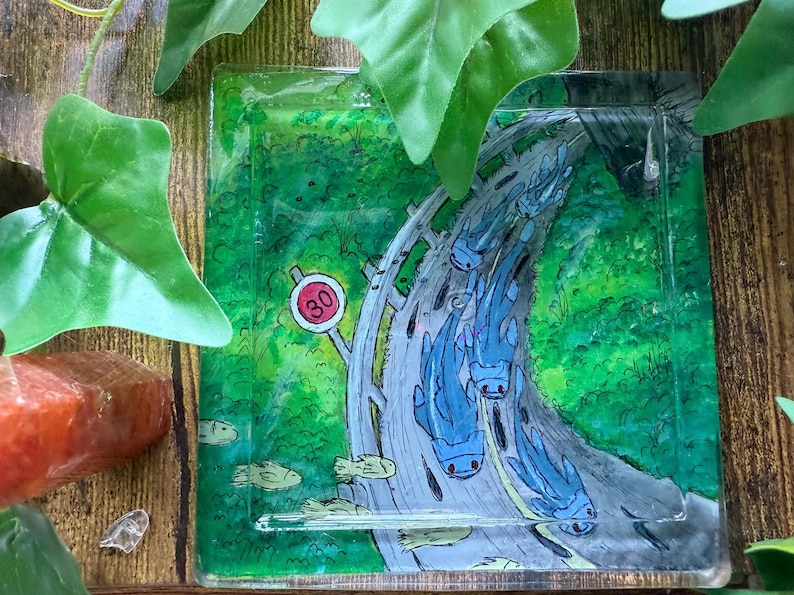 May include: Square, transparent coaster with a whimsical design. The coaster features a road scene with blue creatures and a speed limit sign of 30. The background is a vibrant green, with a wooden surface.