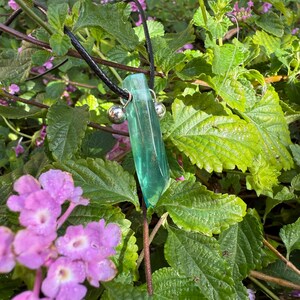 May include: A green crystal pendant necklace with a black cord. The crystal is a long, rectangular shape with a polished surface. Two small silver beads are attached to the crystal. The background features green leaves and purple flowers.