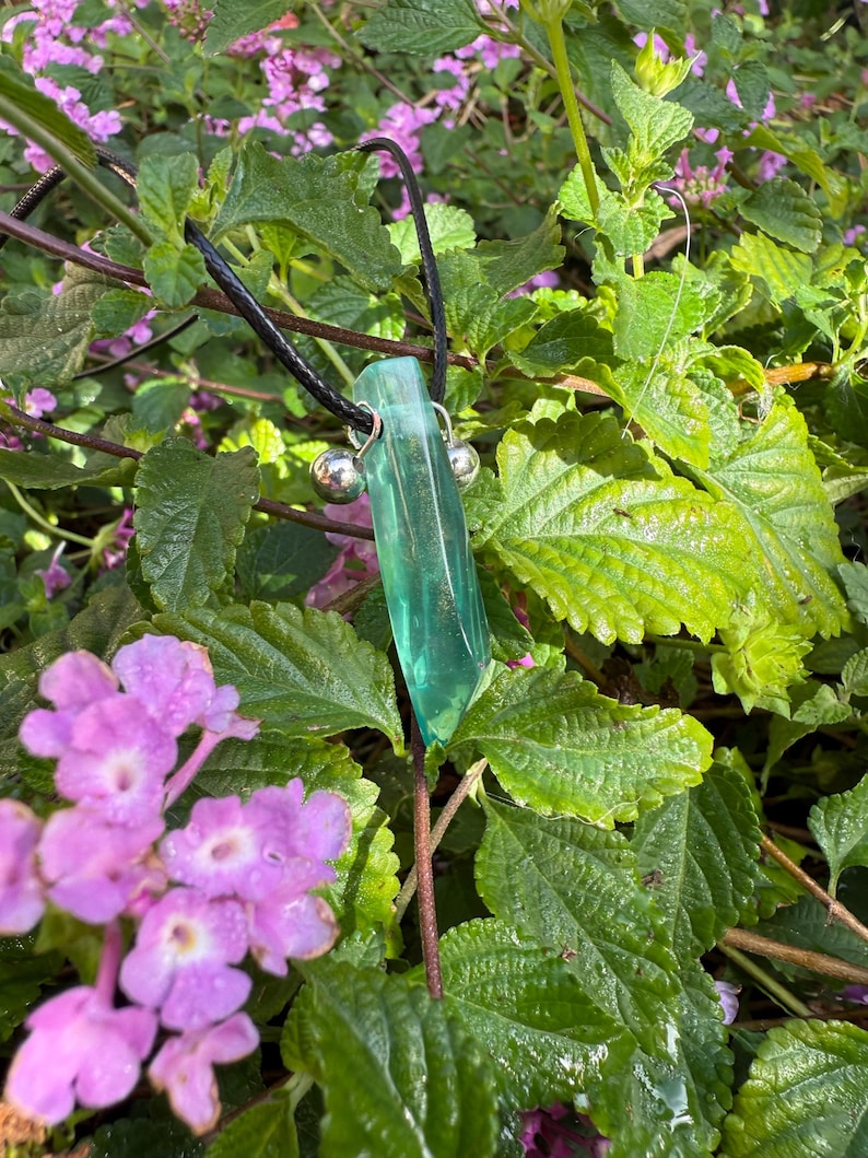May include: A green crystal pendant necklace with a black cord. The crystal is a long, rectangular shape with a polished surface. Two small silver beads are attached to the crystal. The background features green leaves and purple flowers.