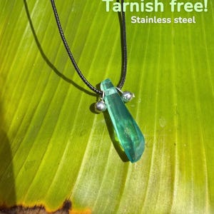 May include: A pendant necklace with a green, crystal-like charm and two small silver beads. The necklace hangs on a black cord against a green leaf. Text on the image reads "Tarnish free! Stainless steel."
