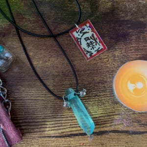 May include: Two necklaces with black cords. One features a rectangular pendant with Japanese characters on a red background. The other has a blue crystal pendant with small silver bells. The items are on a wooden surface.