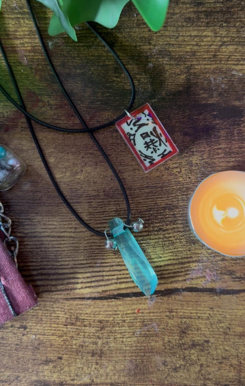 May include: Two necklaces with black cords. One features a rectangular pendant with Japanese characters on a red background. The other has a blue crystal pendant with small silver bells. The items are on a wooden surface.