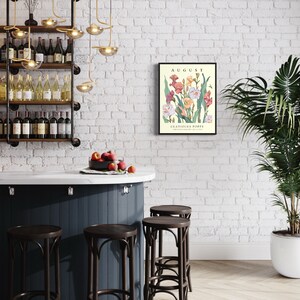 May include: A framed botanical print with a white background and a floral illustration of pink, white, and orange gladiolus flowers. The print is titled "AUGUST" and "GLADIOLUS POPPY". The print is hanging on a white brick wall above a bar counter with four dark brown bar stools.