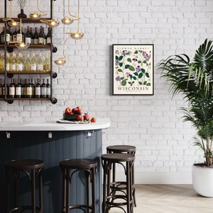 May include: A white brick wall with a black framed print that says "Flower Market Wisconsin" with a botanical illustration of purple flowers. There are four dark brown bar stools in front of a dark blue counter with a white countertop. A bowl of red apples sits on the counter. A potted green plant is in the corner of the room.