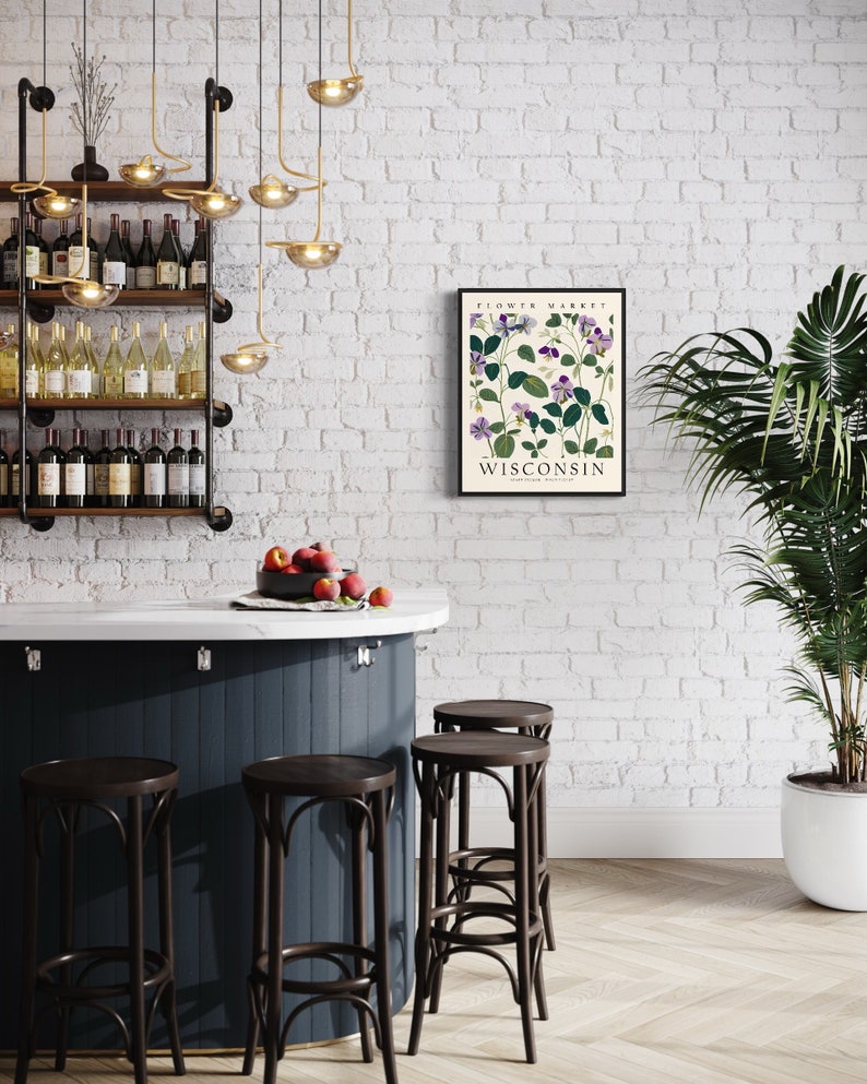 May include: A white brick wall with a black framed print that says "Flower Market Wisconsin" with a botanical illustration of purple flowers. There are four dark brown bar stools in front of a dark blue counter with a white countertop. A bowl of red apples sits on the counter. A potted green plant is in the corner of the room.