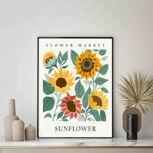 May include: A black framed print with a white background featuring a floral illustration of sunflowers in shades of yellow, orange, and green. The text "FLOWER MARKET" is at the top of the print and "SUNFLOWER" is at the bottom.