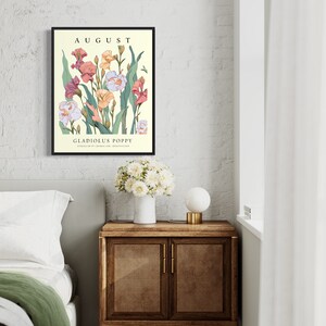 May include: A framed print with a floral illustration of pink, white, and yellow flowers with green leaves. The text "AUGUST" is at the top of the print and "GLADIOLUS POPPY" is at the bottom. The print is hanging on a white brick wall.