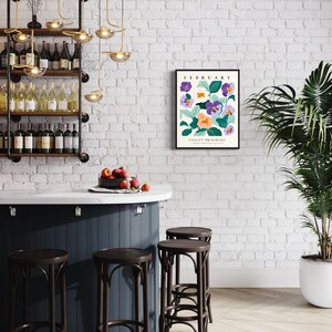 Puede incluir: Una pared de ladrillo blanco con una impresi&oacute;n enmarcada que presenta una ilustraci&oacute;n floral de pr&iacute;mula violeta con el texto "Febrero" y "Pr&iacute;mula violeta" en texto negro. La impresi&oacute;n cuelga sobre una barra de bar azul oscuro con cuatro taburetes de bar marrones. Una planta verde en maceta est&aacute; a la derecha de la imagen.