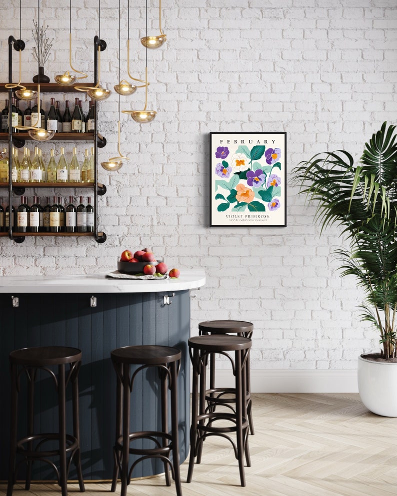Puede incluir: Una pared de ladrillo blanco con una impresi&oacute;n enmarcada que presenta una ilustraci&oacute;n floral de pr&iacute;mula violeta con el texto "Febrero" y "Pr&iacute;mula violeta" en texto negro. La impresi&oacute;n cuelga sobre una barra de bar azul oscuro con cuatro taburetes de bar marrones. Una planta verde en maceta est&aacute; a la derecha de la imagen.