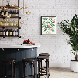 May include: A white brick wall with a black framed print of a botanical illustration of pink flowers with green leaves. The print has the text "FLOWER MARKET IOWA" on it. There is a dark blue bar with four brown bar stools in front of the wall. There are wine bottles on a shelf behind the bar.