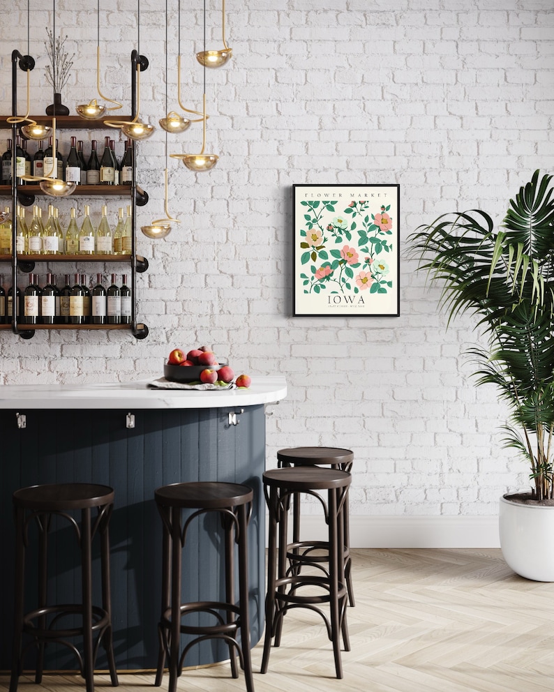 May include: A white brick wall with a black framed print of a botanical illustration of pink flowers with green leaves. The print has the text "FLOWER MARKET IOWA" on it. There is a dark blue bar with four brown bar stools in front of the wall. There are wine bottles on a shelf behind the bar.