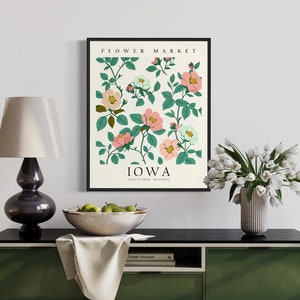 May include: A framed print with a white background featuring a floral illustration of pink and white flowers with green leaves. The text "FLOWER MARKET" and "IOWA" are printed in black at the top and bottom of the print. The print is hanging on a white wall above a black console table with a white tablecloth and a bowl of green apples.