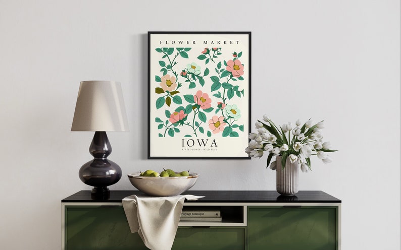 May include: A framed print with a white background featuring a floral illustration of pink and white flowers with green leaves. The text "FLOWER MARKET" and "IOWA" are printed in black at the top and bottom of the print. The print is hanging on a white wall above a black console table with a white tablecloth and a bowl of green apples.