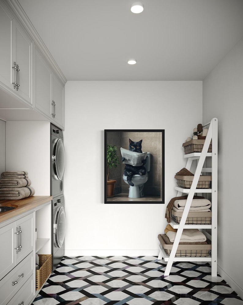 May include: A black cat sitting on a toilet reading a newspaper in a framed picture. The picture is hanging on a white wall in a laundry room with a white tiled floor, a white shelf with baskets, and a washer and dryer.