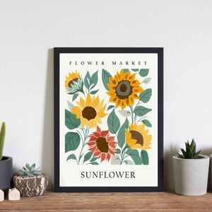May include: A black framed print with a white background featuring a floral illustration of sunflowers in various shades of yellow, orange, and red. The text "FLOWER MARKET" and "SUNFLOWER" are printed in black at the top and bottom of the print.