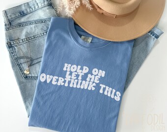 May include: A light blue t-shirt with the text "HOLD ON LET ME OVERTHINK THIS" in white, paired with light wash jeans, a tan hat, and a gold necklace. The shirt is folded and placed on top of the jeans.
