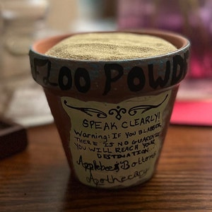 Puede incluir: Una maceta de terracota marrón llena de arena. El borde está pintado con detalles en azul y negro y las palabras "FLOO POWD" están escritas en negro. Una etiqueta dice "SPEAK CLEARLY!" y "Applebee's Bottoms Apothecary."