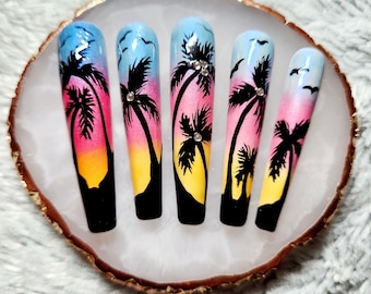May include: Five long, artificial fingernails with a sunset gradient design. Each nail is painted with a different shade of pink, orange, and yellow, and features a silhouette of a palm tree with a blue sky background. The nails are decorated with small, clear rhinestones.