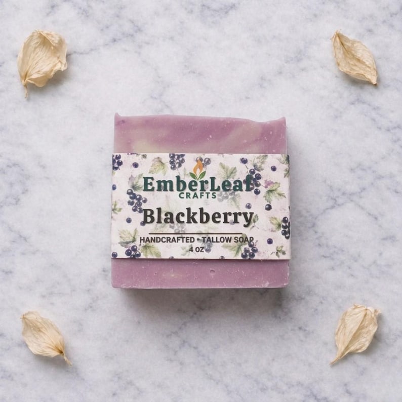 May include: A handcrafted blackberry tallow soap bar, 4 oz, with a purple and white marbled design. The soap is wrapped with a label featuring the text "EmberLeaf Crafts" and "Blackberry" with a blackberry illustration. The soap is surrounded by dried petals.