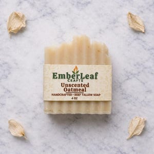 May include: A rectangular bar of unscented oatmeal soap with a paper label. The label reads "EmberLeaf Crafts Unscented Oatmeal Handcrafted Beef Tallow Soap 4 oz." The soap is light beige and sits on a marble surface with dried petals.