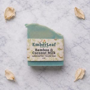 May include: A handcrafted, blue-green tallow soap bar with a label that reads "EmberLeaf Crafts Bamboo & Coconut Milk." The soap is 4 oz. and surrounded by four dried, beige flower petals on a marble surface.