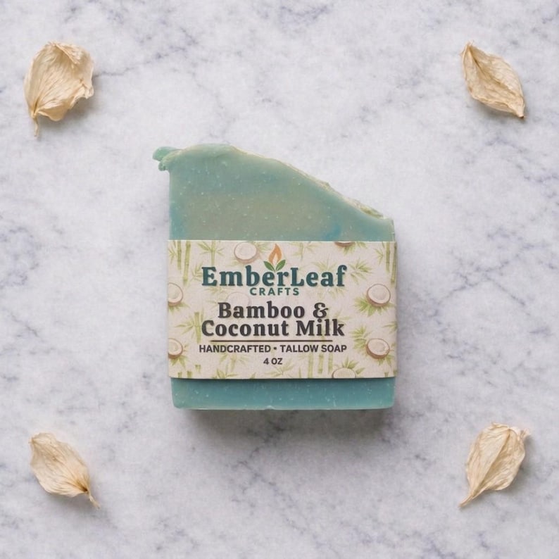 May include: A handcrafted, blue-green tallow soap bar with a label that reads "EmberLeaf Crafts Bamboo & Coconut Milk." The soap is 4 oz. and surrounded by four dried, beige flower petals on a marble surface.