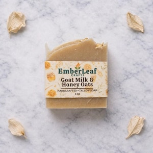 May include: A rectangular bar of Goat Milk & Honey Oats soap from EmberLeaf Crafts, wrapped with a decorative label. The soap is a light beige color, with visible oats. The label includes the text "HANDCRAFTED TALLOW SOAP" and "4 oz".