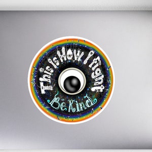 Può includere: Un adesivo rotondo con un bordo arcobaleno e un occhio bianco e nero al centro. Il testo "This is how I fight" è scritto in un cerchio attorno all'occhio, e il testo "Be Kind" è scritto sotto l'occhio.