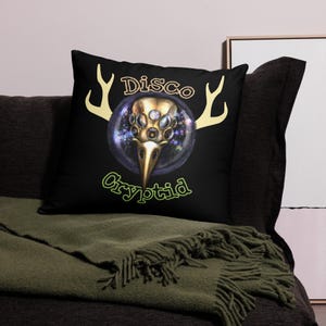 Puede incluir: Cojín negro con un gráfico de una criatura con múltiples ojos y cuernos. El texto "Disco Cryptid" está impreso en la almohada.