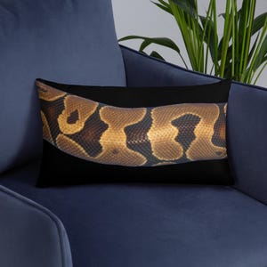 May include: A rectangular pillow with a black background features a close-up of a snake's patterned skin. The snake skin has a mix of brown, tan, and dark brown colors. The pillow is resting on a blue chair.