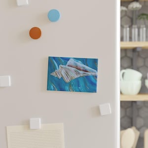 May include: A rectangular magnet featuring a painting of a white seashell centered against a background of swirling blue and gold paint. The magnet is attached to a white surface alongside two small round magnets in blue and orange.