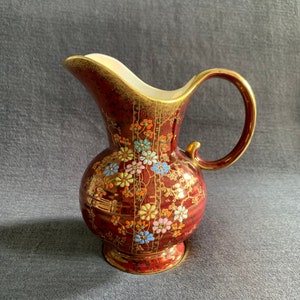 May include: A red and gold ceramic pitcher with a floral design. The pitcher has a round body and a curved handle.