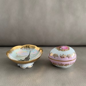 May include: Two small porcelain trinket boxes. One is a round box with a pink and white floral design and gold trim. The other is a shallow bowl with a gold rim and a picture of the Eiffel Tower with the text "Villefranche sur Mer" in gold lettering.