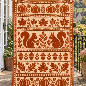 Peut inclure: Une couverture rectangulaire en crochet présentant un motif automnal répétitif en orange brûlé et crème. Le design comprend des rangées de citrouilles, de feuilles d'érable, d'écureuils tenant des glands et de glands isolés, encadrés par une bordure.