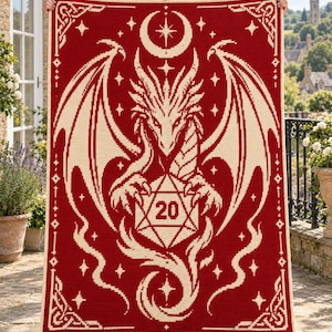 Peut inclure: Une couverture en crochet rectangulaire présentant un fond rouge profond avec un motif couleur crème. Le graphisme central représente un dragon aux ailes déployées tenant un dé à vingt faces avec le chiffre 20, surmonté d'un croissant de lune.