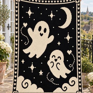 Peut inclure: Une couverture en crochet rectangulaire avec un fond noir et un motif crème représentant deux fantômes, un croissant de lune, des étoiles et des nuages. La bordure présente un motif répétitif de cœurs crème sur fond noir.