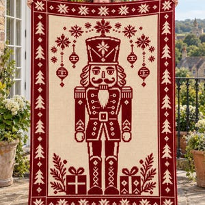 May include: A rectangular knit throw blanket featuring a deep red and cream color scheme. The center displays a large nutcracker figure flanked by two gift boxes. The border is decorated with a repeating pattern of pine trees and snowflakes.