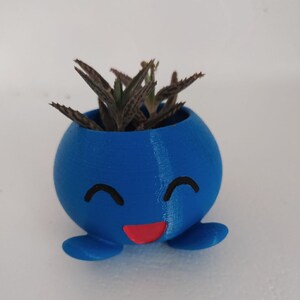 Puede incluir: Una maceta azul impresa en 3D con forma de personaje de dibujos animados con una cara sonriente y una boca roja. Una pequeña planta suculenta está creciendo dentro de la maceta.