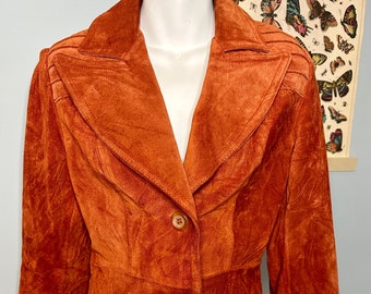 May include: Burnt orange suede blazer with a notched collar, two-button closure, and two front pockets. The blazer has a tailored fit and three-quarter length sleeves. The jacket is displayed on a mannequin.