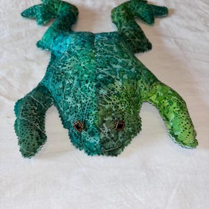 May include: A handmade frog plush toy in shades of green and teal. The frog has button eyes and a quilted pattern on its body. The plush toy is lying on a white surface.