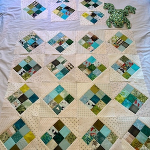 May include: A collection of square quilt blocks featuring a patchwork of teal, green, and white fabric squares arranged in a diamond pattern, accompanied by a plush green frog toy with dark spots and yellow eyes positioned on the upper right side.