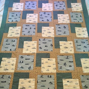 May include: A patchwork quilt featuring alternating square blocks of light blue and cream fabric, each printed with small illustrations of sailboats and lighthouses. The blocks are framed by tan and dark teal fabric borders with a subtle texture.