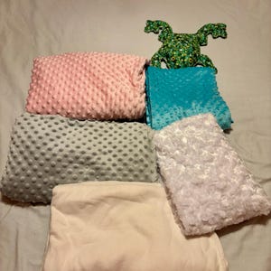 May include: A collection of soft blankets in various colors, including pink, teal, gray, white, and cream. A green frog-shaped plush toy is also present. The blankets have textured surfaces, some with a dot pattern, others with a fluffy texture.