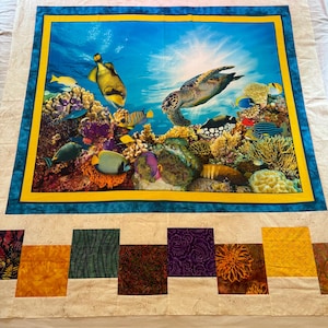 May include: A colorful quilt featuring a vibrant underwater scene with fish, coral, and a sea turtle. The central image is framed with a blue and yellow border. The quilt has a white background with patterned squares in various colors.