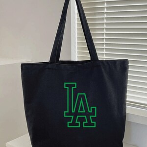 Pode incluir: Bolsa tote de lona preta com um logotipo LA verde.