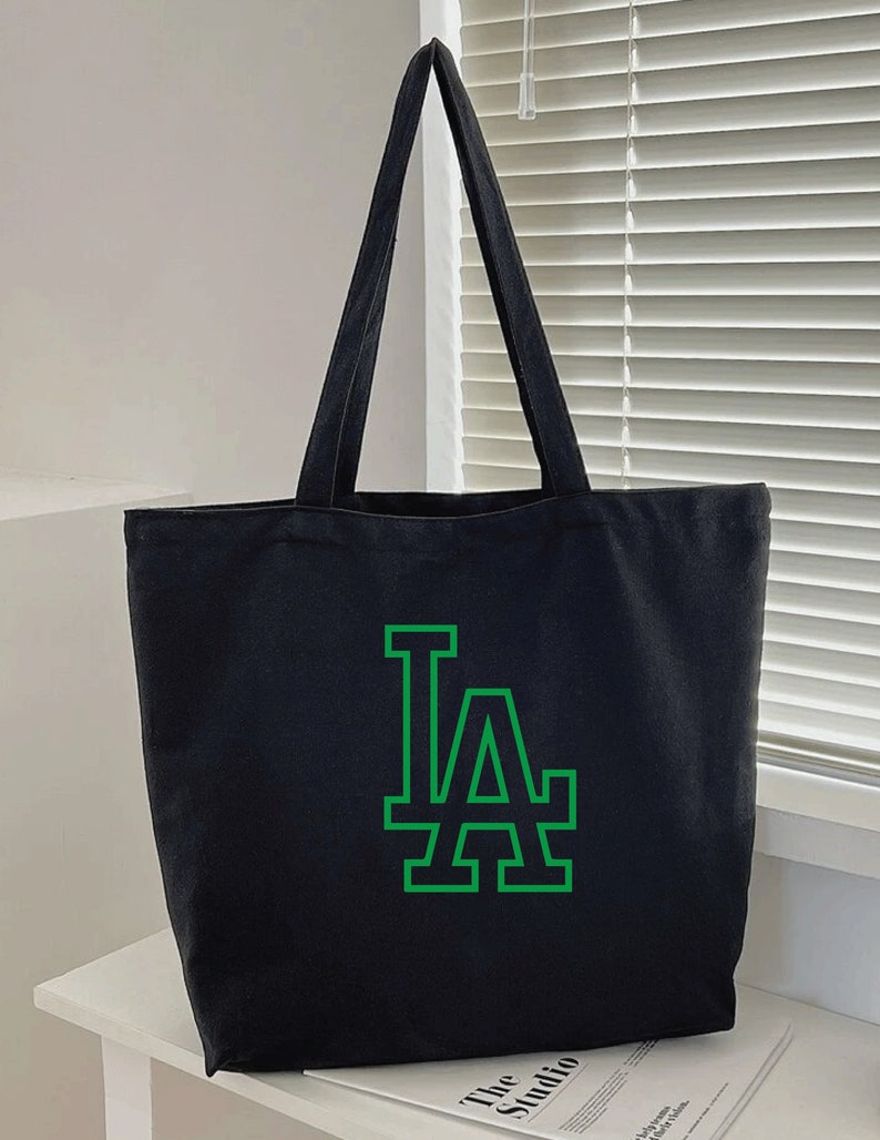 Pode incluir: Bolsa tote de lona preta com um logotipo LA verde.