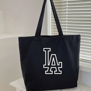 Pode incluir: Sacola de lona preta com um logotipo "LA" branco.