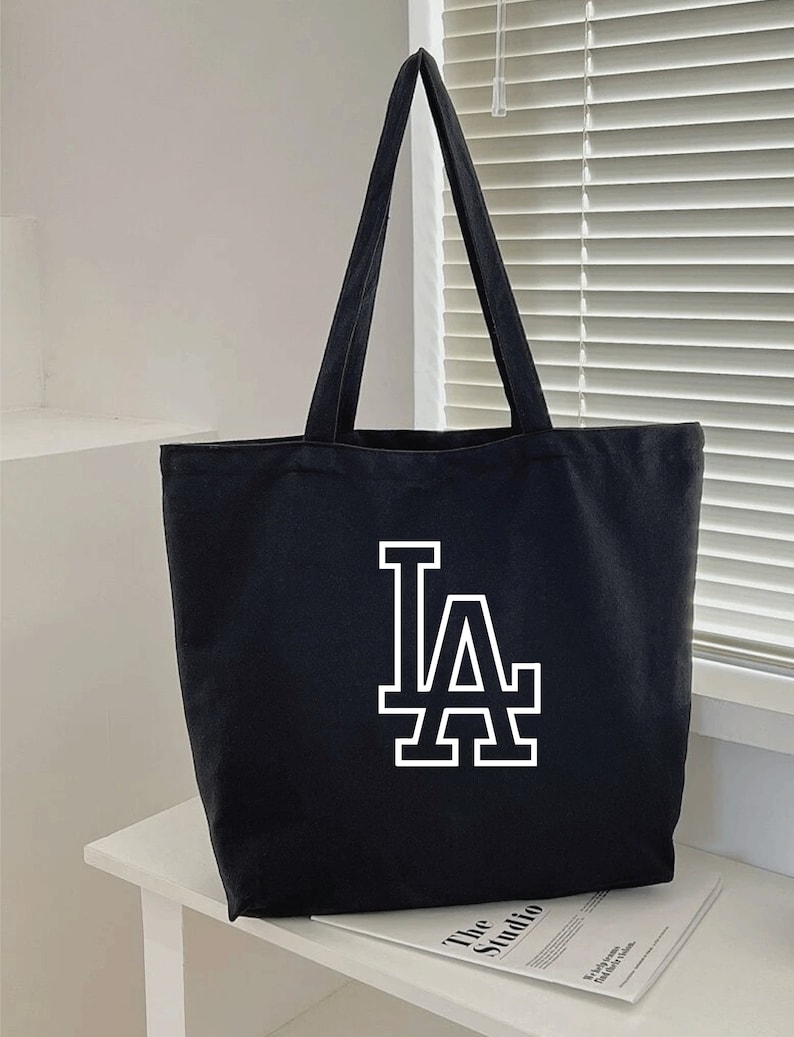 Pode incluir: Sacola de lona preta com um logotipo "LA" branco.