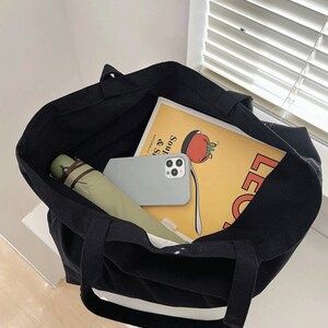 Pode incluir: Uma bolsa tote de lona preta com acabamento branco, cheia de um guarda-chuva verde, uma capa de telefone branca e um livro amarelo com o texto "Soups & Salads" e uma ilustra&ccedil;&atilde;o de tomate vermelho.
