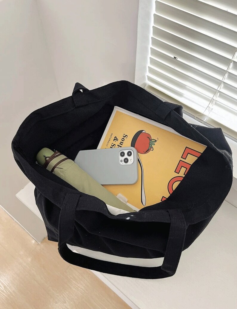 Pode incluir: Uma bolsa tote de lona preta com acabamento branco, cheia de um guarda-chuva verde, uma capa de telefone branca e um livro amarelo com o texto "Soups & Salads" e uma ilustra&ccedil;&atilde;o de tomate vermelho.